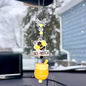 May include: A car charm featuring a bee-themed design. The charm includes a yellow and black bee, the text "BEE-YOTCH", and a honey pot with the word "HUNNY". The charm hangs from a silver chain with a clear bow and yellow beads.