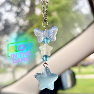 May include: A car charm with a butterfly, star, and bead design. The charm is suspended from a silver chain. The charm has a blue and white color scheme. The text "GLOW IN THE DARK" is visible in the image.