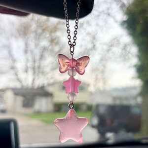May include: A pink car charm with a bow, two stars, and clear beads. The charm hangs from a silver chain. The bow and stars are a translucent pink color. The charm is hanging in front of a blurred background.