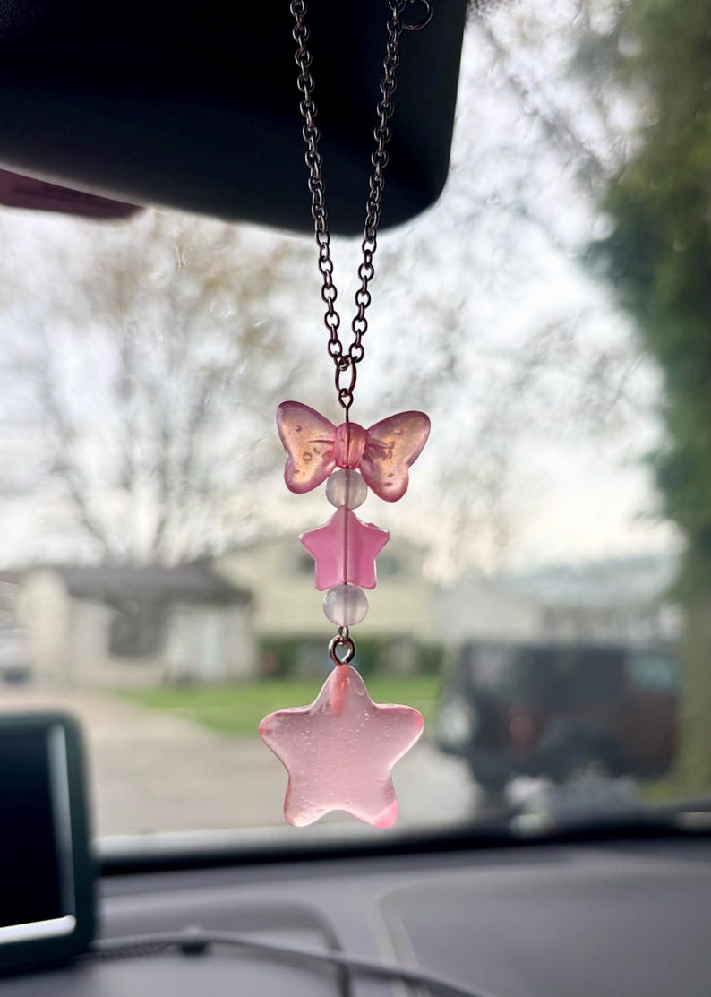May include: A pink car charm with a bow, two stars, and clear beads. The charm hangs from a silver chain. The bow and stars are a translucent pink color. The charm is hanging in front of a blurred background.