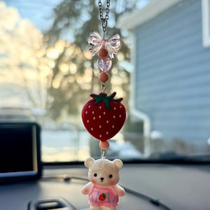 May include: A car charm featuring a red strawberry, a white bear wearing a pink outfit, and a clear bow. The charm hangs from a silver chain and is designed to be hung from a car's rearview mirror.