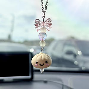May include: A hanging car charm featuring a silver-toned chain, a translucent pink bow bead, a small iridescent heart bead, a round pale bead, and a beige dumpling-shaped charm with a smiling face, black eyes, and pink cheeks.