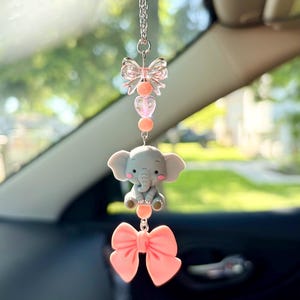 May include: A car charm featuring a gray elephant figurine with pink cheeks, a pink bow, and a clear bow. The charm hangs from a silver chain and includes pink beads and heart-shaped accents. The background is blurred.