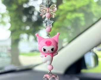 May include: A pink and white car charm featuring a cartoon character with a pink hat and a heart-shaped charm. The charm is suspended from a silver chain, with a clear bow and star accents.