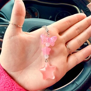 May include: A pink charm with a butterfly, stars, and beads. The charm is suspended from a silver chain. The butterfly and stars are translucent pink. The charm is held in a hand.