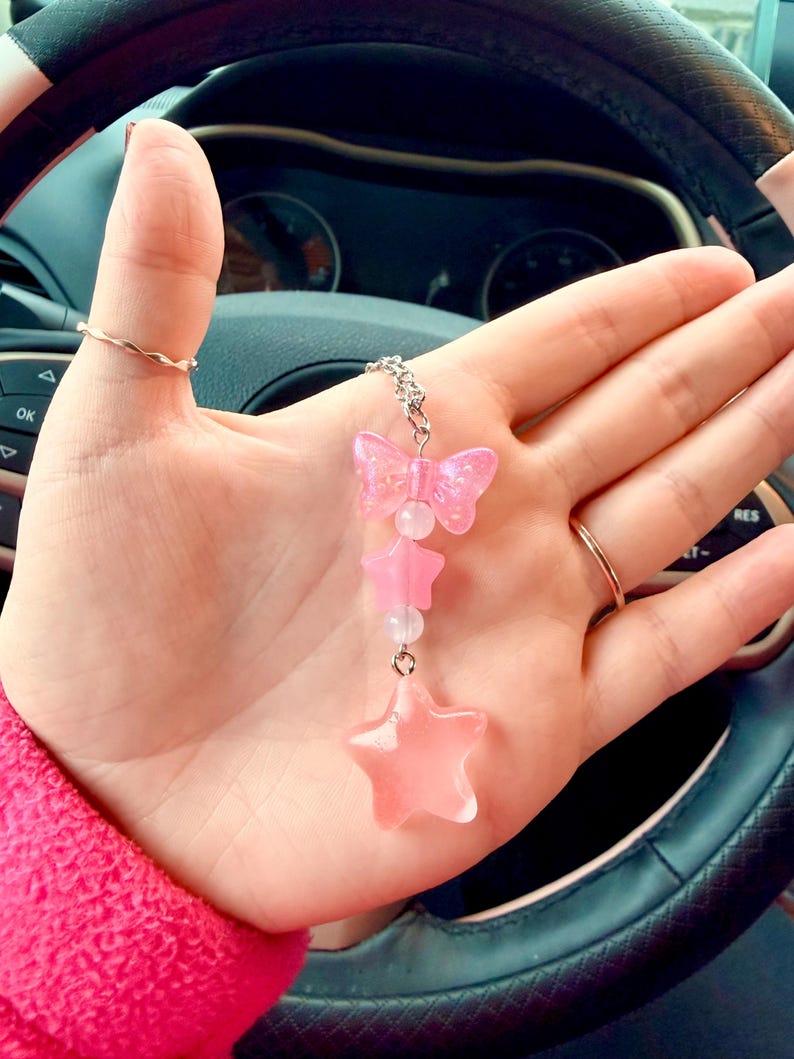 May include: A pink charm with a butterfly, stars, and beads. The charm is suspended from a silver chain. The butterfly and stars are translucent pink. The charm is held in a hand.