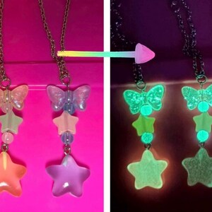 May include: Three necklaces with butterfly and star pendants. The pendants have a gradient of colors, including pink, orange, and purple. The necklaces glow in the dark, with the butterfly and stars emitting a bright green light.