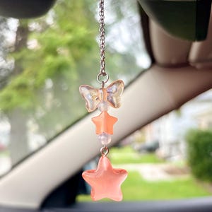 May include: A car charm featuring a silver chain with a butterfly, star, and bead design. The butterfly and stars are a translucent peach color. The charm hangs from a car's rearview mirror.