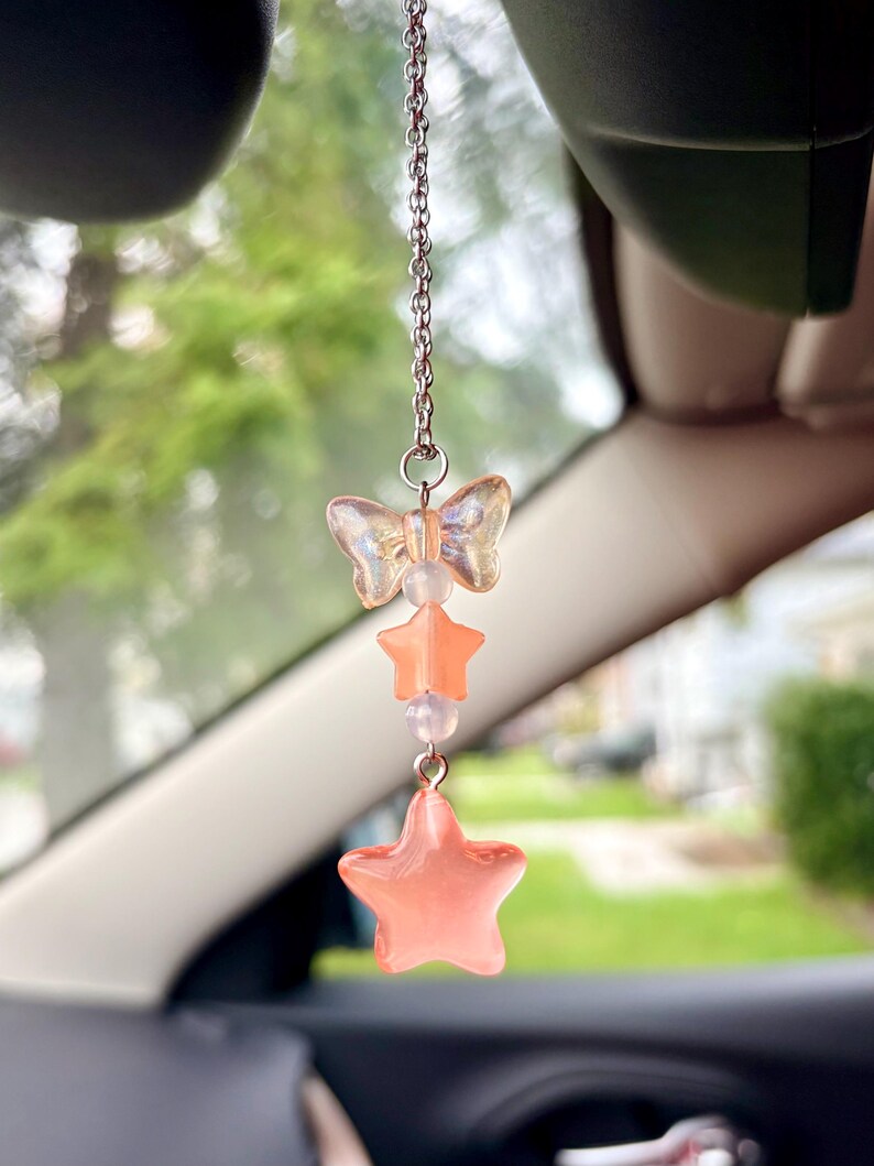 May include: A car charm featuring a silver chain with a butterfly, star, and bead design. The butterfly and stars are a translucent peach color. The charm hangs from a car's rearview mirror.