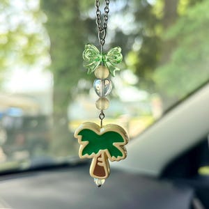 May include: A hanging car charm featuring a silver-toned chain, a translucent light green bow bead, two small clear spherical beads, a heart-shaped bead, and a palm tree pendant with a green canopy and a tan trunk with brown details.