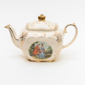 Może przedstawiać: Kremowa porcelanowa czajniczek z złotymi zdobieniami i złotą koroną na pokrywce. Czajniczek posiada malowaną scenę dwóch postaci w strojach z epoki. Klasyczny element dla miłośników herbaty.
