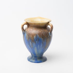 Peut inclure: Un vase en céramique avec une base en piédestal et deux anses. Le vase présente un dégradé de couleurs, passant d'un bord beige clair à des teintes marron et bleues. La couleur bleue semble couler sur les côtés.