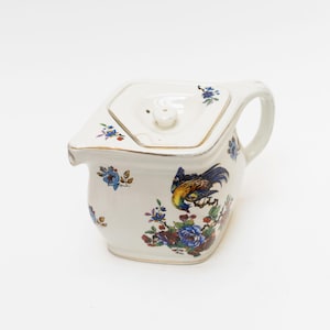 May include: A cream-coloured ceramic creamer with a lid, featuring a gold-trimmed rim and handle. The body is decorated with a colourful illustration of a bird perched on a branch surrounded by blue, red, and purple floral patterns.