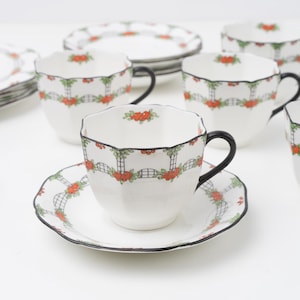 May include: A set of white porcelain teacups and saucers with a scalloped edge, featuring a repeating pattern of black lattice trellises, green leaves, and orange flowers, finished with a thin black rim and a solid black curved handle.
