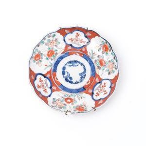 May include: A decorative ceramic plate with a scalloped edge. The plate features a red border with white floral designs and a blue and white center with a detailed pattern. The plate is likely for display or serving.