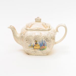 Puede incluir: Tetera de cerámica color crema con tapa y asa. La tetera está decorada con motivos florales dorados y una colorida ilustración de una persona con un vestido amarillo. El pico y el asa son curvos.