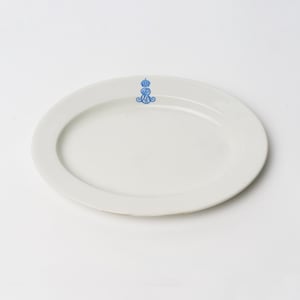 May include: A white, oval-shaped ceramic platter with a blue emblem near the top. The emblem appears to be a crown. The platter is on a white background.
