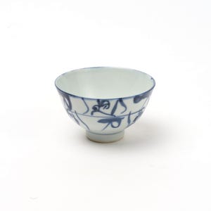 以下が含まれることがあります： 青い花柄の白い陶器のボウル。ボウルは丸みを帯びた形で、小さな足とわずかにフレアした縁があります。青いデザインはボウルの外側にペイントされています。