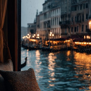 May include: A cozy scene of a rainy day in Venice. A cup of coffee sits on a wooden table next to a lit candle. A textured pillow rests on a window seat, overlooking the canal with gondolas and city lights.