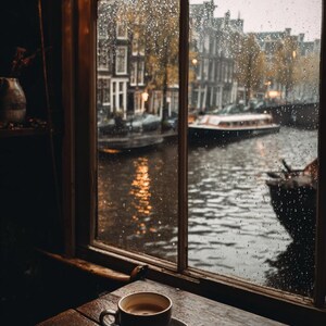 May include: A coffee cup and saucer on a wooden table by a window. Raindrops obscure the view of a canal with buildings and boats. The image creates a cosy, rainy day atmosphere, typical of a European city.