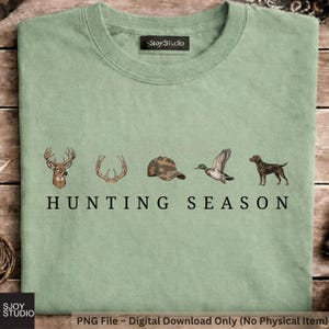May include: A sage green t-shirt featuring a graphic print of a deer head, antlers, a camouflage baseball cap, a flying mallard duck, and a brown hunting dog. Below the illustrations, the text reads HUNTING SEASON in black capital letters.