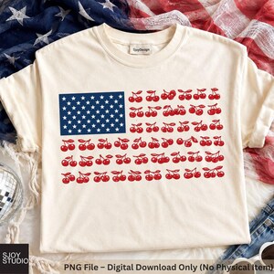 Puede incluir: Una camiseta de color crema con un diseño gráfico de la bandera estadounidense donde las franjas están reemplazadas por filas de cerezas rojas. El cantón azul contiene estrellas blancas. La etiqueta del cuello muestra el texto SjoyDesign.