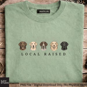 Pode incluir: Uma t-shirt verde sálvia com um gráfico de cinco raças de cães diferentes dispostas numa fila horizontal. Abaixo dos cães, o texto LOCAL RAISED aparece numa fonte serifada preta. A t-shirt tem gola redonda e uma etiqueta SjoyStudio.