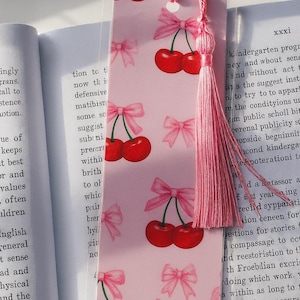 May include: A pink bookmark featuring a pattern of cherries and bows. The bookmark has a pink tassel attached. The cherries are red, and the bows are pink. The bookmark is rectangular and rests on an open book.