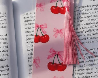 Può includere: Segnalibro rosa con motivo a ciliegie e fiocchi. Il segnalibro ha un nappa rosa attaccata. Le ciliegie sono rosse e i fiocchi rosa. Il segnalibro rettangolare è posto su un libro aperto.