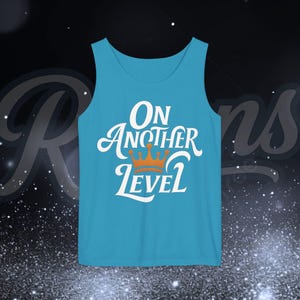 May include: A turquoise tank top with the phrase "On Another Level" in white script and a gold crown graphic. The text is centered on the front of the shirt. The background is dark with a subtle pattern.