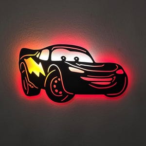 May include: Black Lightning McQueen car-shaped wall art with a red neon glow around the edges. The car has a yellow lightning bolt design and two round headlights. A fun, decorative piece for a child's room or a car enthusiast.