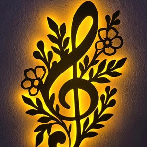 Puede incluir: Una pieza de arte mural de metal negro con una clave de sol, iluminada con un cálido resplandor amarillo. El símbolo musical está rodeado de acentos florales y de hojas, creando un diseño decorativo y artístico. La obra de arte mural está montada en una pared gris oscuro.