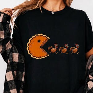 May include: A black t-shirt featuring a graphic of a large orange pumpkin pie slice shaped like a video game character, with three brown turkeys running in a line toward the open mouth of the pie slice, mimicking a classic arcade game scene.