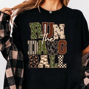 May include: A black t-shirt featuring the text RUN THE DANG BALL in large block letters. Each letter has a unique pattern including football plays, helmets, stripes, goal posts, footballs, and foam fingers in shades of green, brown, and white.