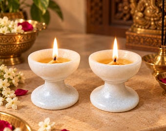 May include: Two white marble oil lamps with a smooth, rounded bowl shape and a sturdy pedestal base. Each lamp features a lit wick centred in a pool of oil, casting a warm glow. The stone has a subtle, speckled texture throughout the surface.
