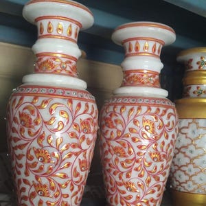 K&ouml;nnte beinhalten: Zwei Vasen aus wei&szlig;em Marmor mit ausgestelltem Rand und zylindrischem Hals. Die Oberfl&auml;che ist mit komplizierten roten und goldenen Blumenrankenmustern verziert, die mit kleinen, auf dem K&ouml;rper und Hals eingebetteten Kristallen akzentuiert sind.