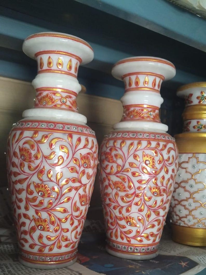 K&ouml;nnte beinhalten: Zwei Vasen aus wei&szlig;em Marmor mit ausgestelltem Rand und zylindrischem Hals. Die Oberfl&auml;che ist mit komplizierten roten und goldenen Blumenrankenmustern verziert, die mit kleinen, auf dem K&ouml;rper und Hals eingebetteten Kristallen akzentuiert sind.