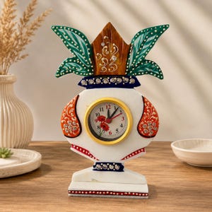 Puede incluir: Un reloj de mesa decorativo con forma de kalash, que presenta una base blanca con detalles en naranja, una parte superior en forma de hojas verdes y pedrería incrustada. La esfera redonda tiene un borde dorado, manecillas negras y flores.
