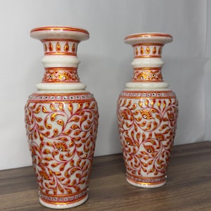 K&ouml;nnte beinhalten: Zwei Vasen aus wei&szlig;em Marmor mit handgemalten Blumenmustern in Rot und Gold. Jede Vase hat einen ausgestellten Rand, einen schlanken Hals und einen bauchigen K&ouml;rper, der mit kleinen, in das detaillierte florale Design eingebetteten Kristallen verziert ist.