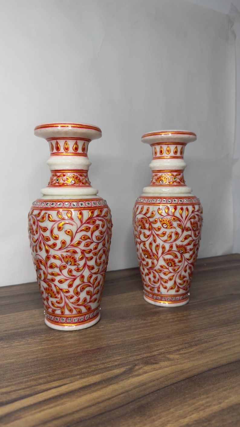 K&ouml;nnte beinhalten: Zwei Vasen aus wei&szlig;em Marmor mit handgemalten Blumenmustern in Rot und Gold. Jede Vase hat einen ausgestellten Rand, einen schlanken Hals und einen bauchigen K&ouml;rper, der mit kleinen, in das detaillierte florale Design eingebetteten Kristallen verziert ist.