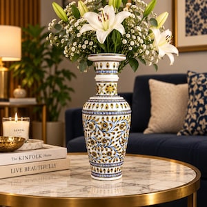 K&ouml;nnte beinhalten: Eine hohe, wei&szlig;e Keramikvase mit einem komplexen, handbemalten Muster aus blauen und gelben Blumenmotiven und tropfenf&ouml;rmigen Elementen. Die Vase hat einen ausgestellten Rand, einen schlanken Hals und einen bauchigen K&ouml;rper, gef&uuml;llt mit wei&szlig;en Lilien.
