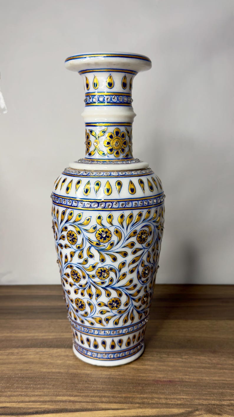 K&ouml;nnte beinhalten: Eine Vase aus wei&szlig;em Marmor mit handgemalten floralen Mustern in Gold und Blau. Die Oberfl&auml;che ist mit kleinen, eingelassenen klaren Kristallen verziert, die in B&auml;ndern und in den Mitten der gemalten Blumen angeordnet sind und ein Dekor bilden.