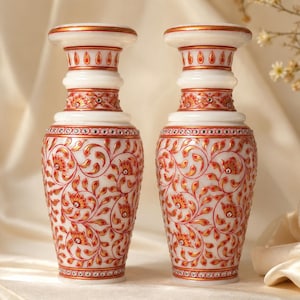 K&ouml;nnte beinhalten: Zwei Vasen aus wei&szlig;em Marmor mit handgemalten Blumenmustern in Rot und Gold. Jede Vase hat einen ausgestellten Rand, einen schlanken Hals und einen bauchigen K&ouml;rper, der mit kleinen eingelegten Kristallen am Hals und Fu&szlig; verziert ist.