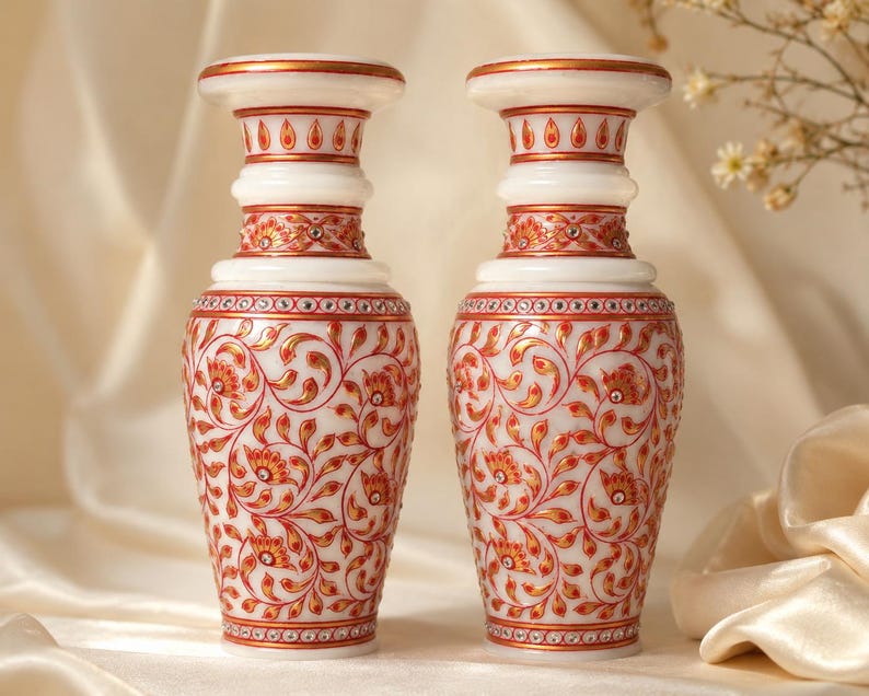 K&ouml;nnte beinhalten: Zwei Vasen aus wei&szlig;em Marmor mit handgemalten Blumenmustern in Rot und Gold. Jede Vase hat einen ausgestellten Rand, einen schlanken Hals und einen bauchigen K&ouml;rper, der mit kleinen eingelegten Kristallen am Hals und Fu&szlig; verziert ist.