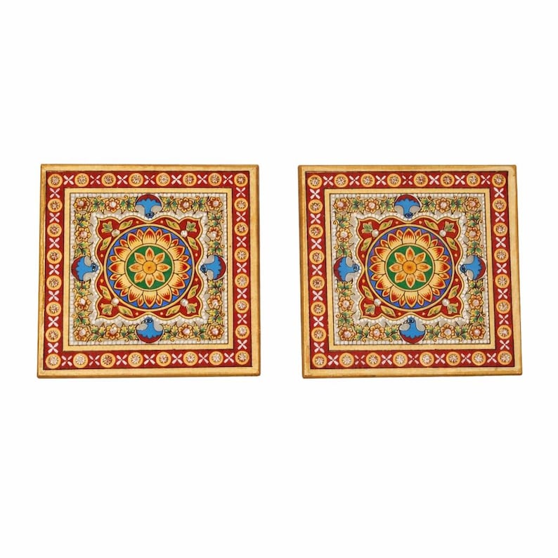 May include: Two square coasters featuring a central floral mandala design in yellow, green, and blue, surrounded by a red border with white and gold geometric patterns and small blue bird motifs, finished with a gold-toned outer edge.