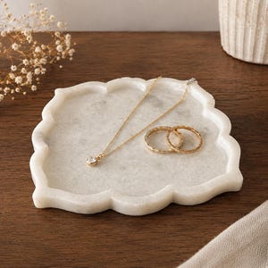 Könnte beinhalten: Ein weißes Marmortablett mit gewelltem Rand hält eine goldene Halskette mit einem einzelnen Anhänger und zwei goldenen Ringen. Das Tablett steht auf einer dunklen Holzoberfläche, mit einer kleinen Vase und Trockenblumen im Hintergrund.