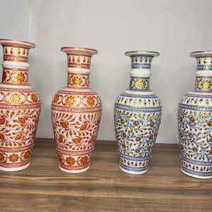 K&ouml;nnte beinhalten: Vier wei&szlig;e Marmorvasen mit schmalen H&auml;lsen und ausgestellten R&auml;ndern. Zwei Vasen zeigen komplizierte Blumenmuster in Rot und Gold, w&auml;hrend die anderen beiden &auml;hnliche florale Designs in Blau und Gold aufweisen. Jede Vase hat einen zylindrischen K&ouml;rper.
