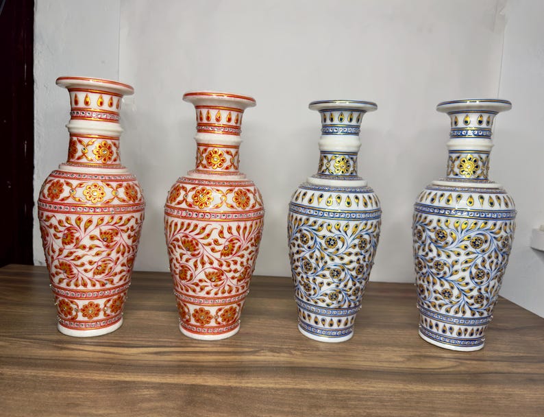 K&ouml;nnte beinhalten: Vier wei&szlig;e Marmorvasen mit schmalen H&auml;lsen und ausgestellten R&auml;ndern. Zwei Vasen zeigen komplizierte Blumenmuster in Rot und Gold, w&auml;hrend die anderen beiden &auml;hnliche florale Designs in Blau und Gold aufweisen. Jede Vase hat einen zylindrischen K&ouml;rper.