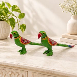 May include: Two green parrot figurines with red beaks and long tails. Each bird features a decorative red wing panel with colorful floral patterns and a yellow collar detail. The parrots are standing side by side, showcasing a vibrant hand-painted finish.
