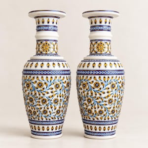 K&ouml;nnte beinhalten: Ein Paar hohe, wei&szlig;e Keramikvasen mit aufwendigen, handbemalten Blumenmustern in Gelb-, Blau- und Goldt&ouml;nen. Das Design umfasst sich wiederholende Blattmotive, kleine Bl&uuml;ten sowie geometrische Bord&uuml;ren am Hals und am Fu&szlig; der Vasen.